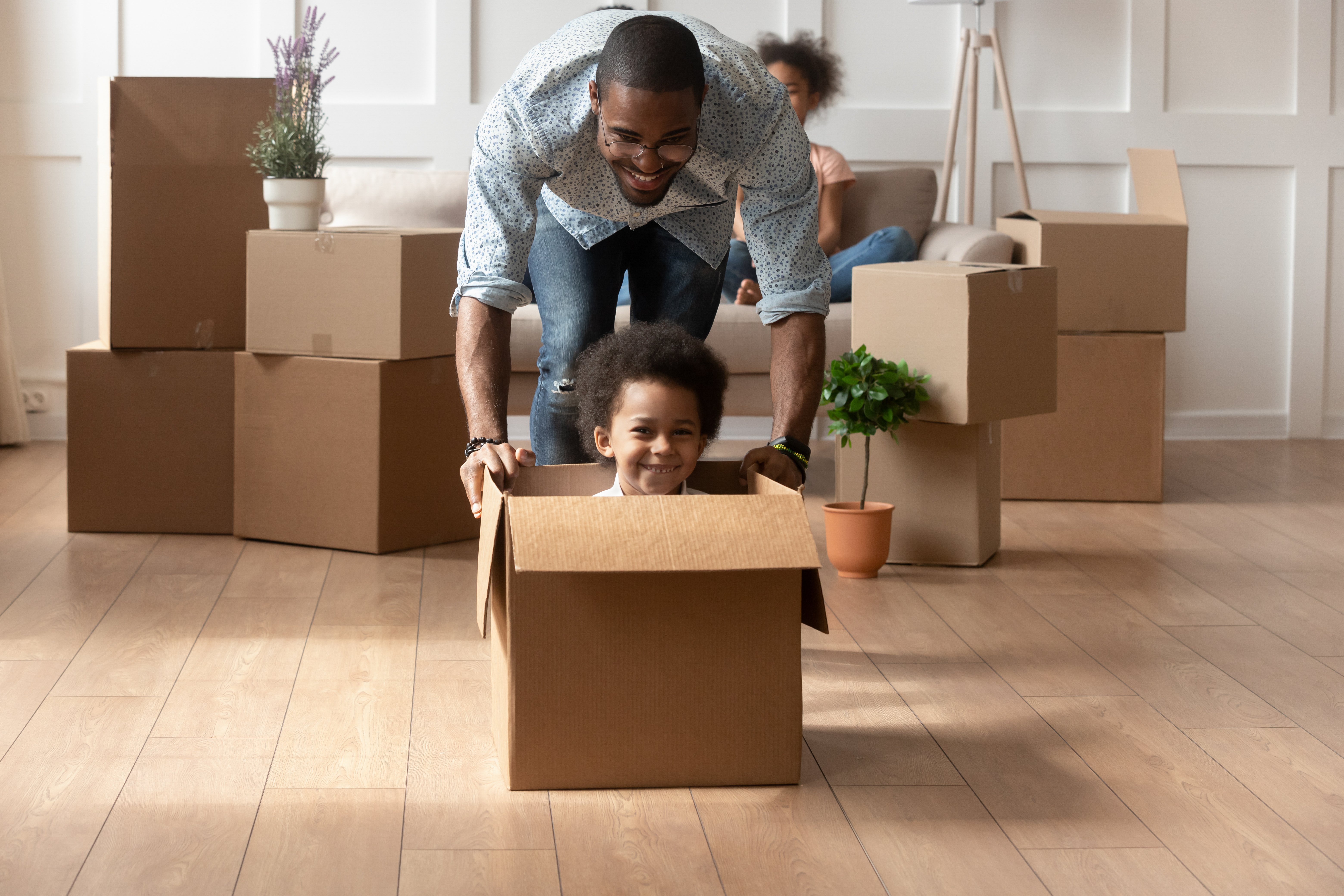 Happy african dad playing with cute little mixed race son looking at camera laughing on moving day, black father having fun with kid boy riding in box in living room, relocation, new house purchase