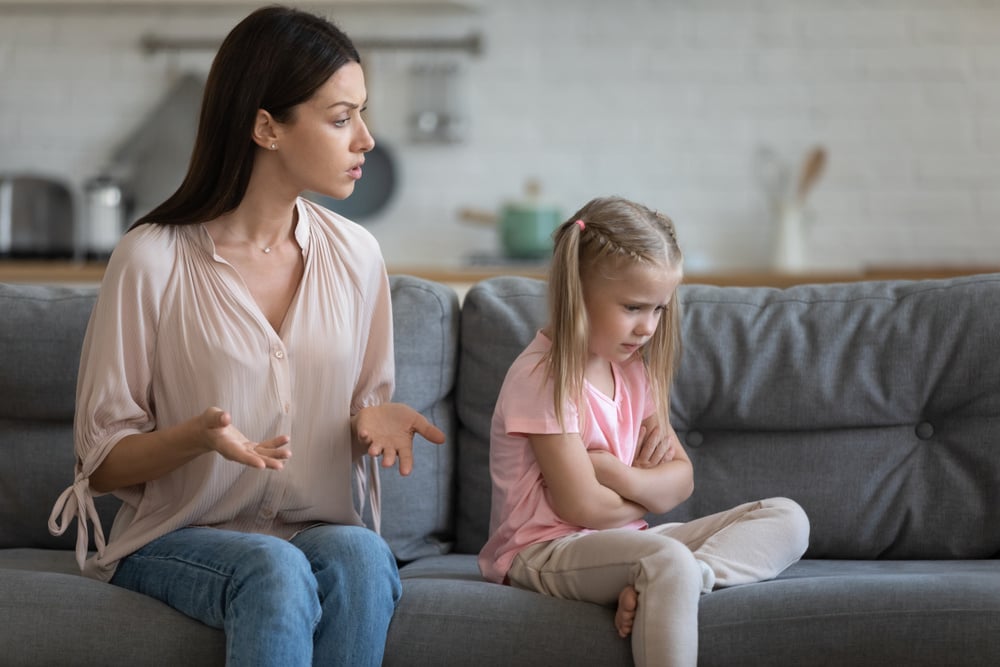 Stubborn upset little daughter ignoring strict young mother, sitting with arms crossed on couch in living room