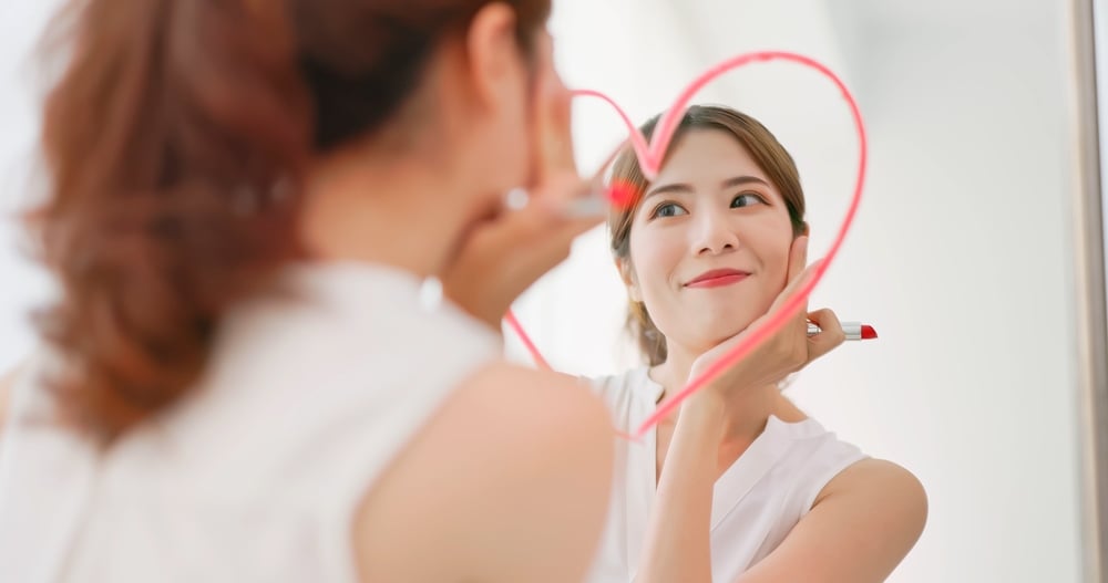 asian woman with brunette ponytail draws a heart shape use lipstick on the mirror and look at herself feeling confident self love