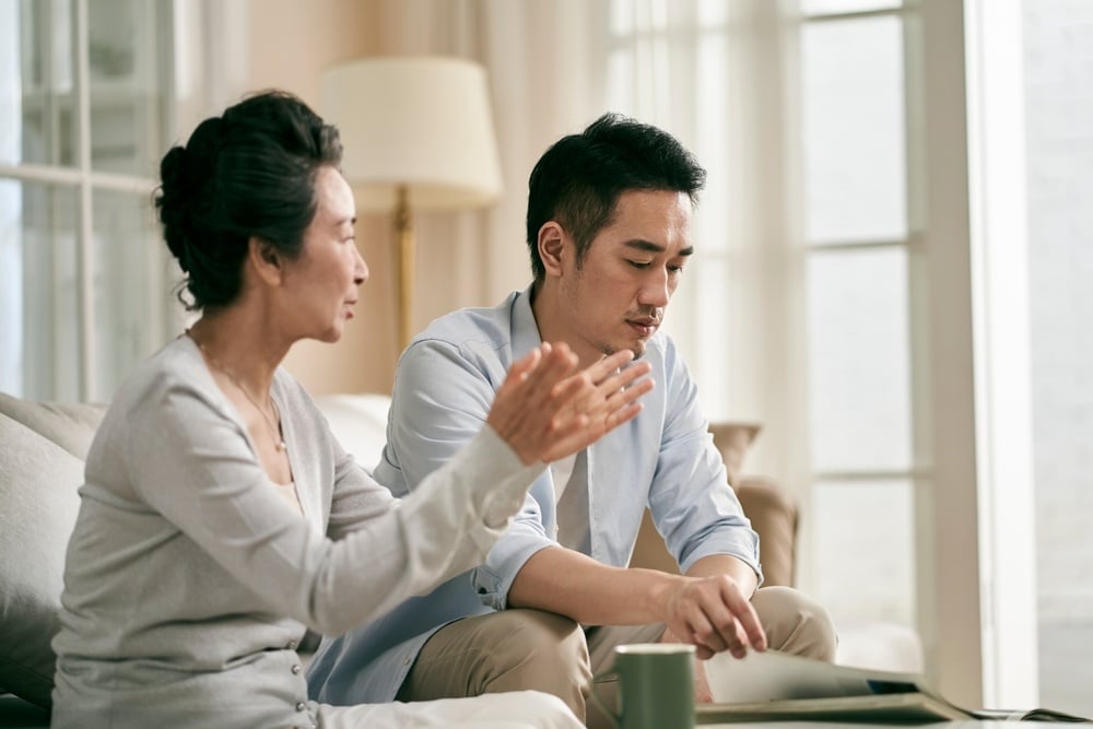 senior asian mother and adult son sitting on couch in living room at home having a conversation