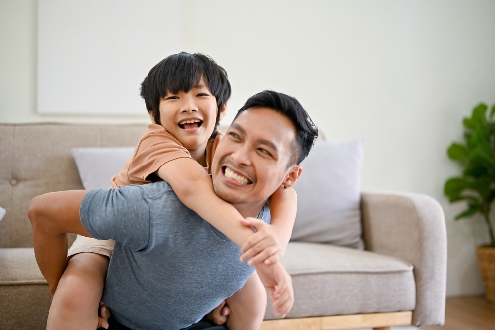 Joyful and happy Asian single dad playing with his little son in the living room, piggyback, spending fun time together at home.