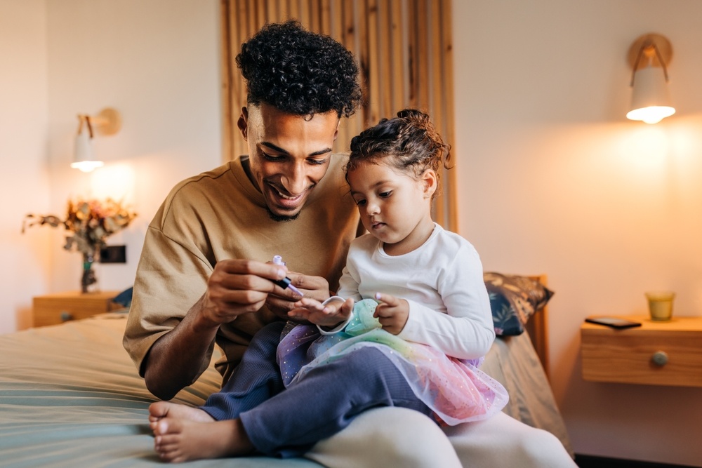 Smiling single dad painting his daughter's nails with nail polish while sitting on a bed. Happy young father spending some quality time with his adorable daughter at home.