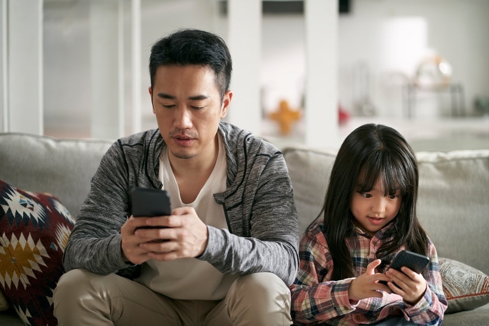 young asian father and seven-year-old daughter sitting on family couch using cellphone 