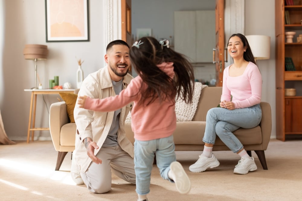 Asian Kid Daughter Running To Embrace Her Dad, While Mother Sitting On Sofa In Modern Living Room