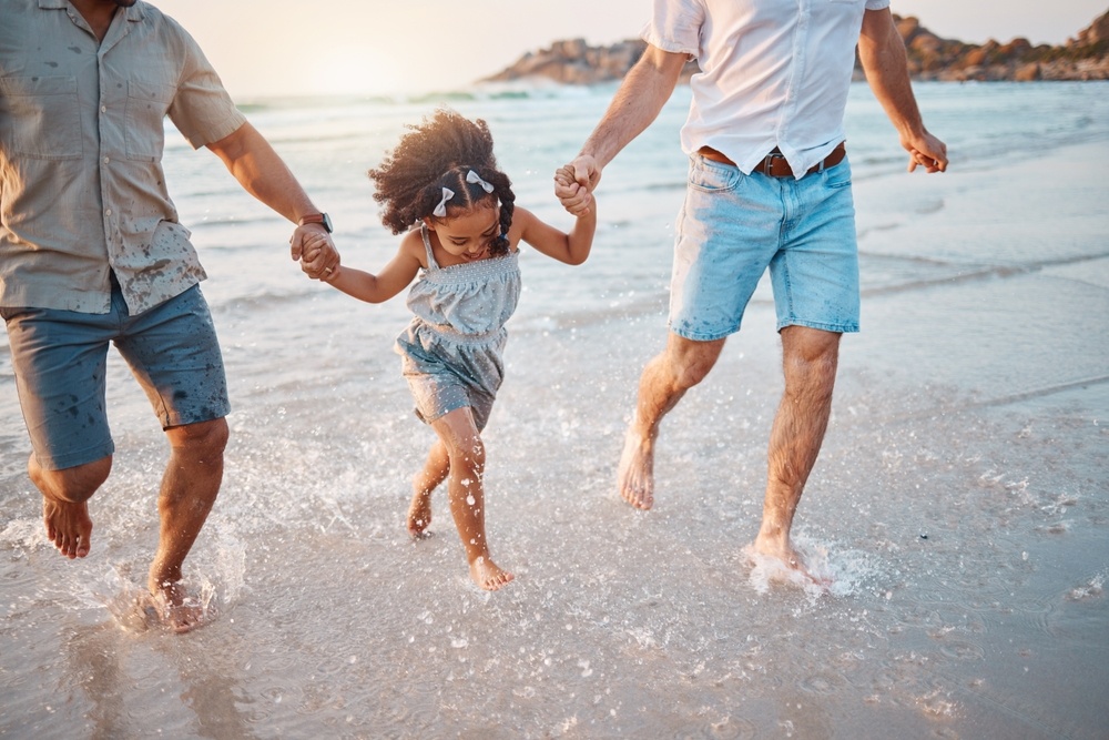 Happy family running on beach with two LGBTQ+ parents holding their daughter's hands between them