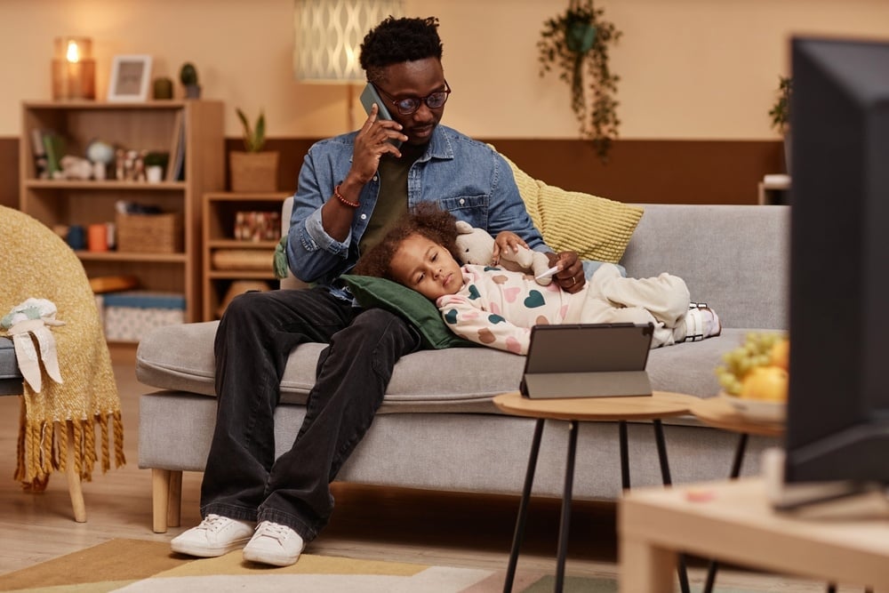 Wide shot of sick African American girl lying on couch and hugging toy looking at tablet while her concerned father talking on phone