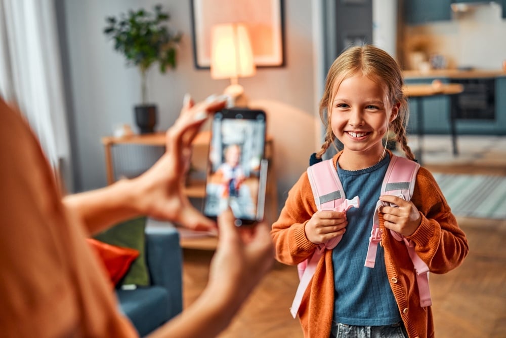 Female holding cellular phone for photographing preteen child getting ready for school.The girl stands in the living room with a backpack and poses for a photo for her mother.